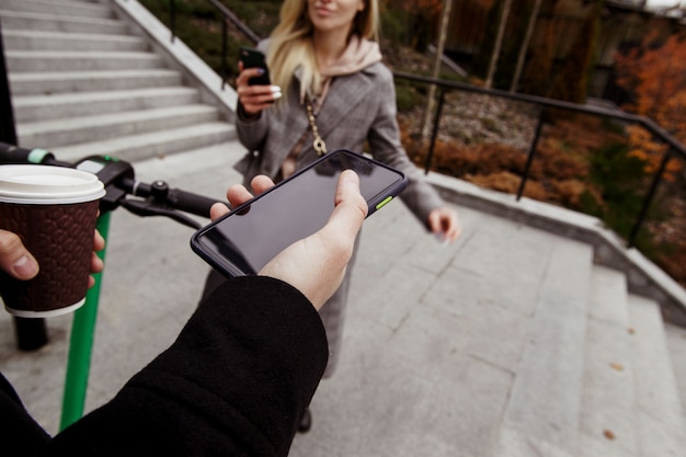 Encontro romântico incomum em scooters elétricos. visão em primeira pessoa. homem segurando smartphone com tela de espaço de cópia em branco, xícara de café e roda. mulher em pé perto com o telefone na mão e sorrindo.