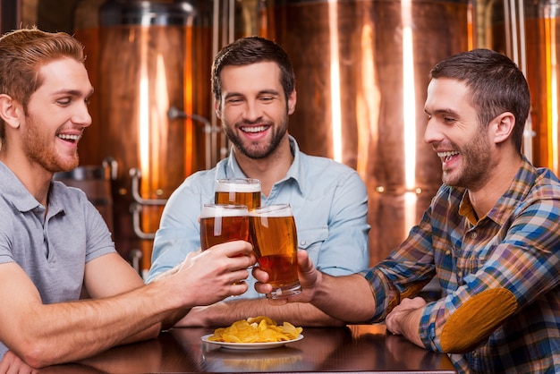 Encuentro con viejos amigos en el bar. Tres hombres jóvenes felices en ropa casual brindando con cerveza y sonriendo mientras están sentados juntos en el pub de cerveza