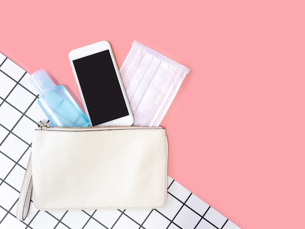 Foto endecha plana de bolso de mujer de cuero blanco con teléfono inteligente, máscara quirúrgica y desinfectante de gel de alcohol sobre fondo rosa, vista superior con espacio de copia de texto.