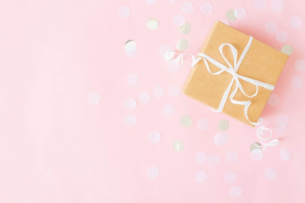 Endecha plana con caja de regalo de papel artesanal aislada atada con cinta, confeti de papel de estrella y círculo o brillos en una superficie rosa pastel