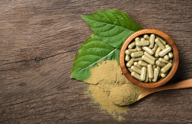 Endecha plana de cápsulas de Kratom (Mitragyna speciosa) con polvo y hojas frescas sobre fondo de madera.