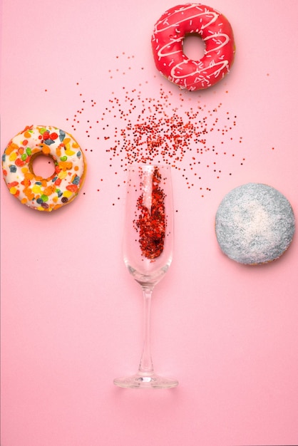 Endecha plana de celebración. Copa de champán con serpentinas de fiesta de colores y deliciosas donas sobre fondo rosa.