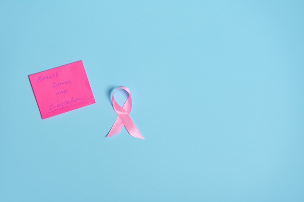 Endecha plana de cinta rosa con un extremo sin fin y una nota de papel con letras 1 de octubre Conciencia sobre el cáncer de mama, aislado sobre fondo azul con espacio de copia. Conceptos médicos y de atención de la salud de la mujer.