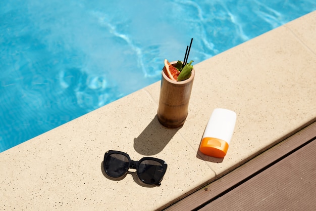 Endecha plana de cóctel en recipiente de madera, gafas de sol negras de moda y crema de protección solar en botella blanca, que compone la composición para descansar durante las vacaciones. Las cosas están situadas cerca de la piscina.