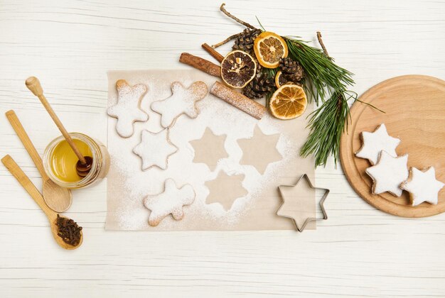 Endecha plana de galletas de jengibre horneadas en forma de estrella espolvoreadas con azúcar para Navidad sobre papel pergamino para hornear cerca de conos de pino y ramitas, rodajas de naranja secas y palitos de canela. De cerca