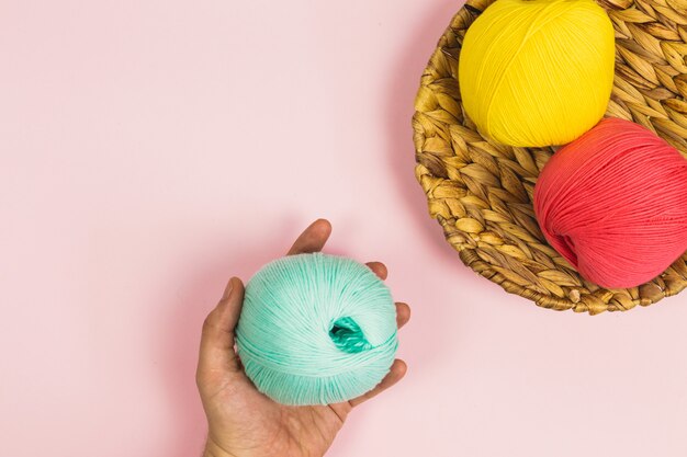 Endecha plana de mano de mujer sosteniendo una hermosa bola de algodón verde menta junto a bolas de algodón de color rosa coral y amarillo oscuro en una canasta con fondo de color rosa pastel y espacio de copia