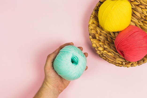 Endecha plana de mano de mujer sosteniendo una hermosa bola de algodón verde menta junto a bolas de algodón de color rosa coral y amarillo oscuro en una canasta con fondo de color rosa pastel y espacio de copia