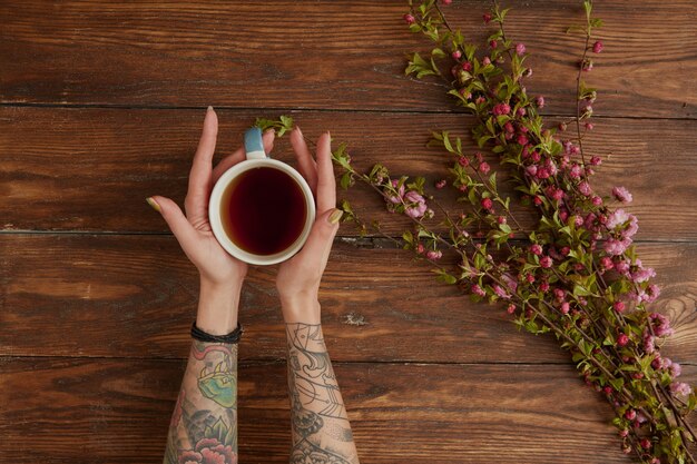 Endecha plana. Mujer sosteniendo una bebida caliente en taza sobre un escritorio de madera con ramas en flor