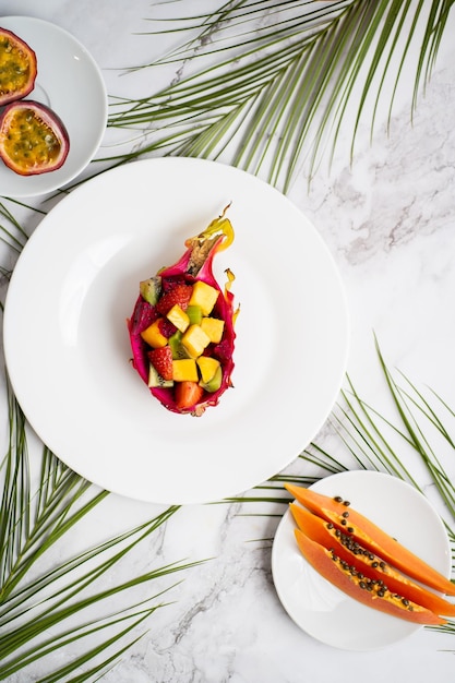 Foto endecha plana de platos con frutas mixtas y frutas en rodajas. ensalada de frutas tropicales servida en media fruta del dragón. fruta del dragón, maracuyá, papaya, hojas de palma en la mesa. vista superior.