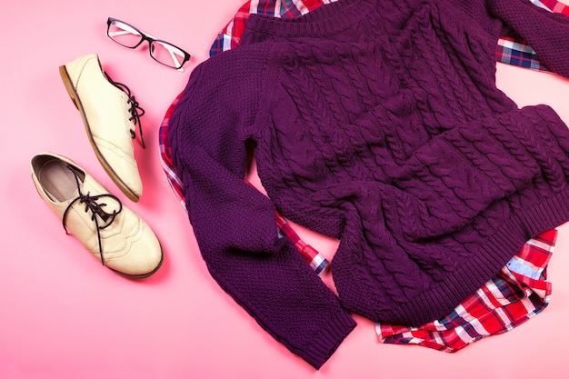 Foto endecha plana de ropa de mujer y accesorios con suéter morado, camisa a cuadros, gafas, botas. fondo rosa