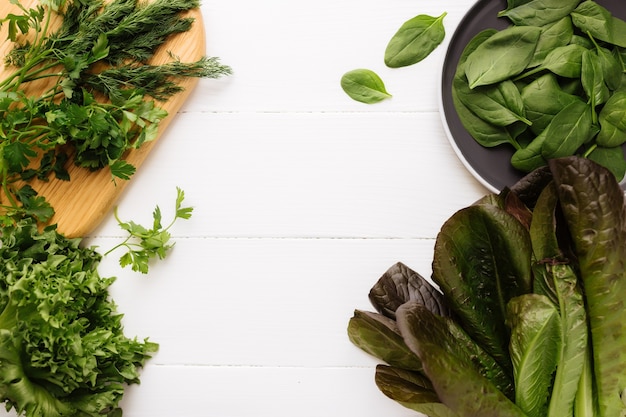 Endecha plana con tabla de cortar y tazón de fuente de ensalada verde fresca hojas de espinaca y lechuga, lechuga romana y perejil, albahaca sobre fondo blanco. Concepto de alimentación saludable