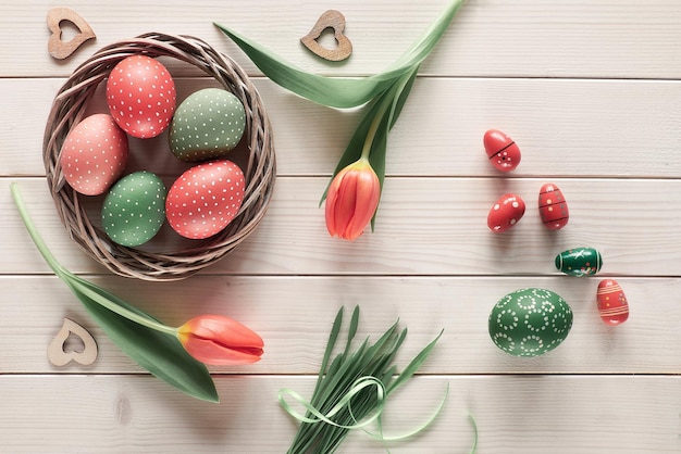 Endecha plana con tulipanes rojos en cesta de corona de mimbre y hierba verde Huevos de Pascua pintados decorativos Fondo de primavera o Pascua en mesa rústica de madera blanca Cero desperdicio decoración sostenible de bajo impacto