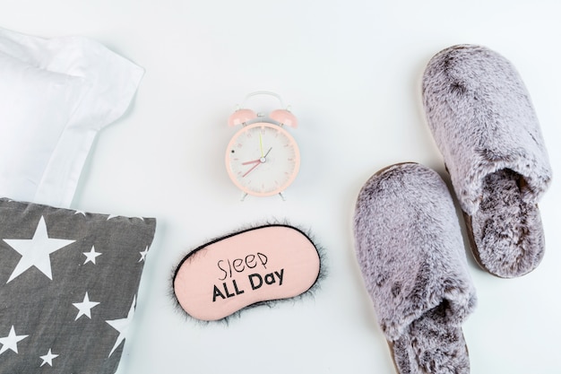 Foto endecha plana con zapatillas suaves y esponjosas, almohada, venda y reloj despertador en blanco