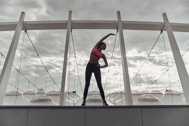 Foto energía en el interior. longitud total de mujer joven moderna en ropa deportiva que se extiende mientras se calienta al aire libre