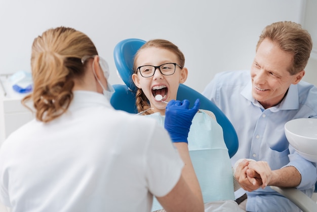 Enérgica adolescente brilhante sentada na cadeira enquanto o médico verifica seus dentes e o pai segura sua mão