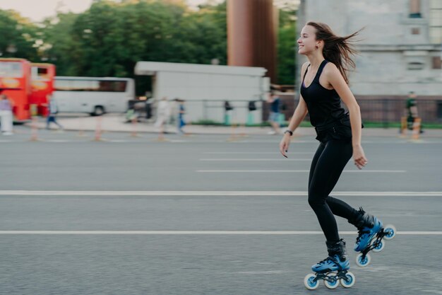 Enérgica, delgada, joven y sonriente mujer patines en la carretera de la ciudad disfruta pasar el tiempo libre se mueve activamente rápidamente tiene el cabello oscuro flotando en el viento lleva un estilo de vida saludable Fines de semana activos Foto al aire libre