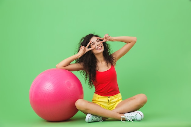 Enérgica mujer vistiendo ropa de verano sentada en el suelo con pelota de fitness durante los ejercicios aeróbicos contra la pared verde