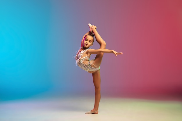 Energie. Kleines kaukasisches Mädchen, rhythmisches Gymnastiktraining, isoliert auf blau-rotem Studiohintergrund mit Farbverlauf in Neon. Anmutig und flexibel, starkes Kind. Konzept von Sport, Bewegung, Aktion.