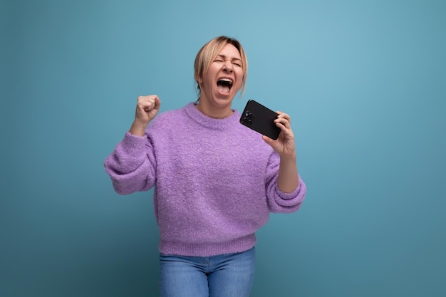 Energiegeladene blonde junge Beraterin in lila Hoodie mit Smartphone mit Mockup und Gesang