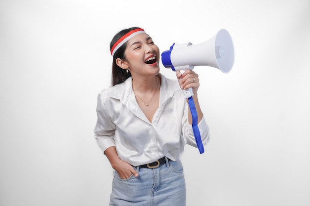 Energische asiatische Frau in formeller Kleidung, die ein Landfahnen-Kopfband trägt, während sie auf einem isolierten weißen Hintergrund an einem Megafon steht und schreit