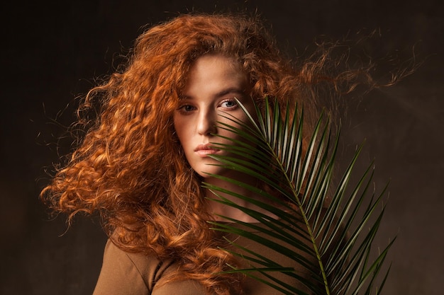 Energizer hermosa mujer joven de cabello rizado jengibre con rama de palma de planta tropical verde posada en estudio de paredes oscuras Fondo del espacio de copia