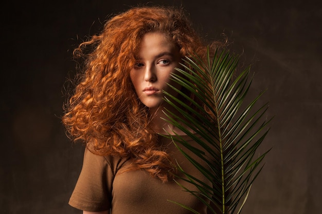 Energizer hermosa mujer joven de cabello rizado jengibre con rama de palma de planta tropical verde posada en estudio de paredes oscuras Fondo del espacio de copia