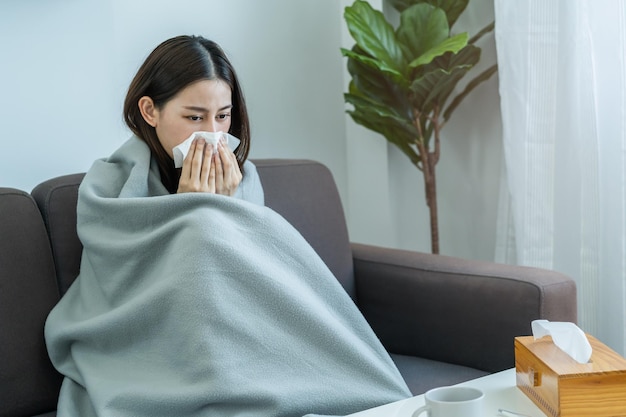 Enferma, Coronavirus covid-19 mujer joven asiática, niña con dolor de cabeza bajo la manta tiene fiebre, gripe y usa pañuelos de papel estornudando la nariz, mocosa sentada en el sofá cama en casa. Atención médica en personas con virus.