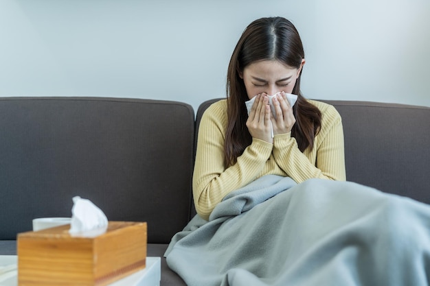 Enferma, Coronavirus covid-19 mujer joven asiática, niña con dolor de cabeza bajo la manta tiene fiebre, gripe y usa pañuelos de papel estornudando la nariz, mocosa sentada en el sofá cama en casa. Atención médica en personas con virus.
