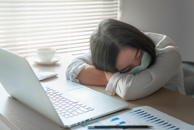 Enferma joven empleada con mascarilla para dormir en el escritorio con computadora portátil