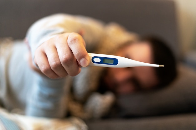 Enfermedad. Anímate en casa. Un joven está enfermo, recibe tratamiento en casa. Medición de la temperatura corporal. En manos de un hombre hay un termómetro electrónico.