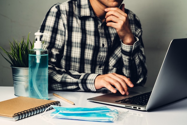 Enfermedad por coronavirus o concepto de protección COVID-19: el hombre joven trabaja en la oficina en casa con equipo de protección y limpieza para protegerse contra el virus de la corona mientras usa una computadora portátil en el escritorio