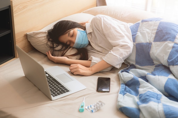 Enfermedad enferma adolescente de la muchacha asiática yacía en la cama con mascarilla con ordenador portátil.