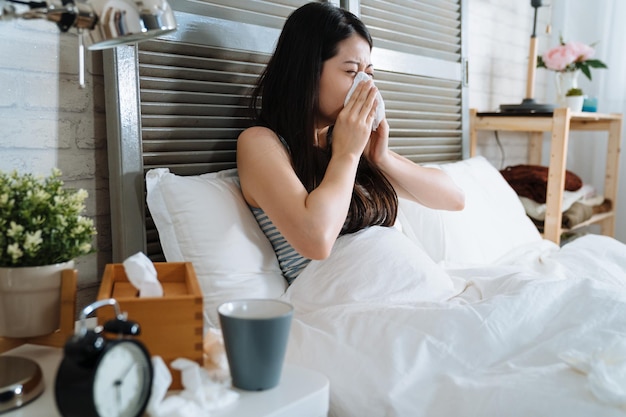 enfermedad Mujer japonesa asiática que se siente mal y estornuda en la cama. hembra que sufre de polilla cubierta fría y nariz con tejido. niña enferma frunciendo el ceño y relajándose en el dormitorio de la luz del sol por la mañana