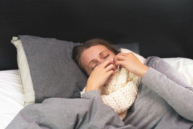 Enfermedad. Tratamiento a domicilio. Una mujer está enferma en casa, moqueo nasal y gripe. Cálidamente vestido y cubierto con una manta.