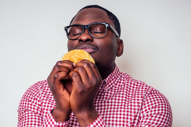 Enfermedad triste hombre afroamericano puso la mano sobre el abdomen de dolor de hamburger.Hombre afro guapo y joven en una elegante camisa y gafas sosteniendo una hamburguesa sobre un fondo blanco. indigestión de la dieta de comida chatarra.