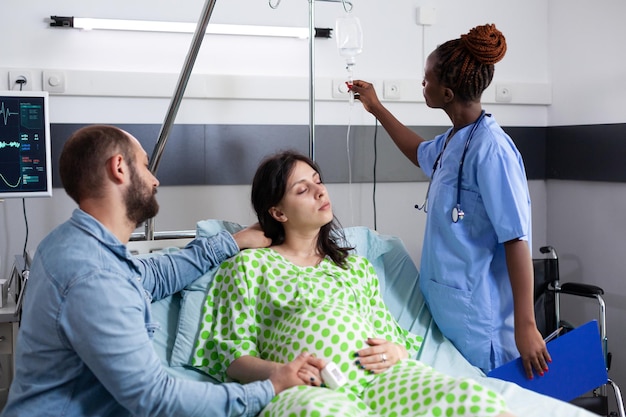 Enfermeira afro-americana verificando gotejamento iv de mulher grávida, preparando-se para cirurgia médica na enfermaria do hospital. Paciente com gravidez deitada na cama, discutindo com o marido sobre paternidade