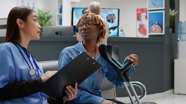 Enfermeira asiática consultando mulher afro-americana com muletas, sentada na área de recepção do hospital. assistente médico e paciente falando sobre deficiência física no saguão da sala de espera.