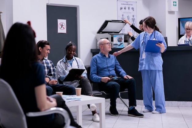 Enfermeira atenciosa levando o paciente sênior para consultar o médico para consulta clínica enquanto o médico afro-americano está preenchendo o formulário para o paciente asiático. Diversas pessoas esperando na recepção do hospital moderno.