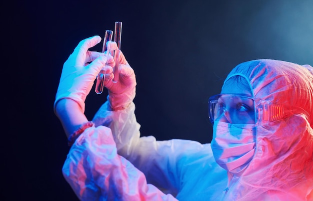 Foto enfermeira de máscara e uniforme branco em pé na sala iluminada de neon e segurando tubos com amostras