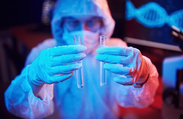 Enfermeira de máscara e uniforme branco segurando tubo com líquido e sentado em laboratório iluminado por neon com computador e equipamento médico procurando vacina coronavirus