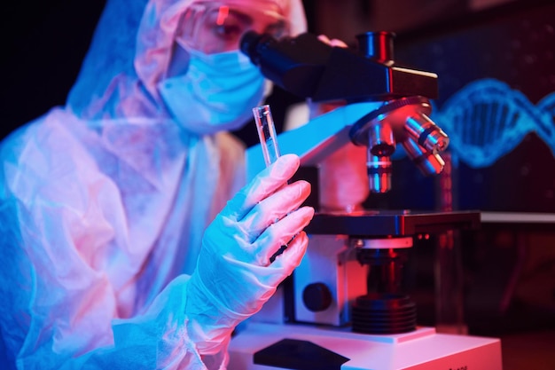 Enfermeira de máscara e uniforme branco segurando tubo com líquido e sentado em laboratório iluminado por neon com computador e equipamento médico procurando vacina Coronavirus