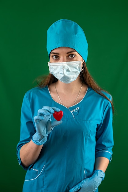 Enfermeira de uniforme azul com luvas de máscara segurando um pequeno coração vermelho na sala de cirurgia do hospital