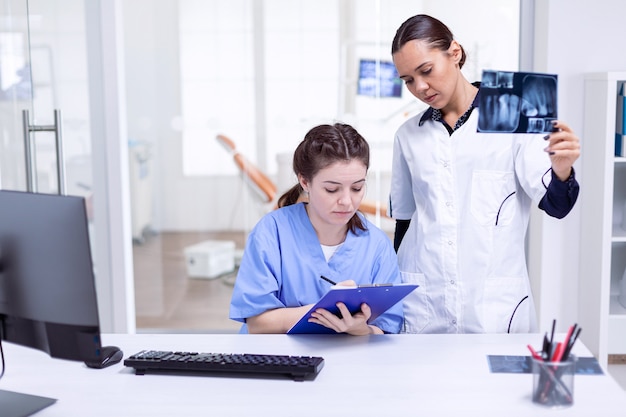 Enfermeira dentista tomando notas sobre a higiene dos dentes enquanto o médico está segurando o raio-x. Stomatolog e seu assistente na recepção da clínica odontológica, olhando para a radiografia de dentes.