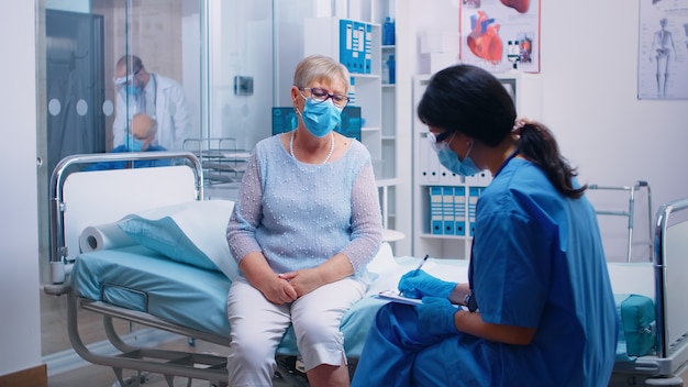 Enfermeira em equipamentos de proteção questionando paciente idosa que usa uma máscara. Hospital, clínica ou centro de saúde privado moderno. Consulta médica durante surto de coronavírus