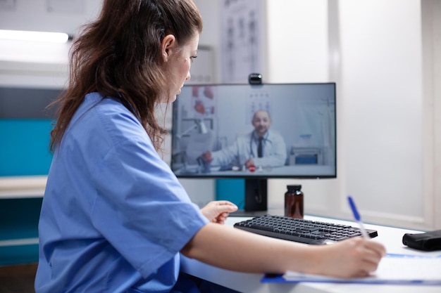 Foto enfermeira em videochamada tomando notas do colega médico em consultório médico profissional. mulher anotando informações recebidas do clínico geral por videoconferência de telemedicina