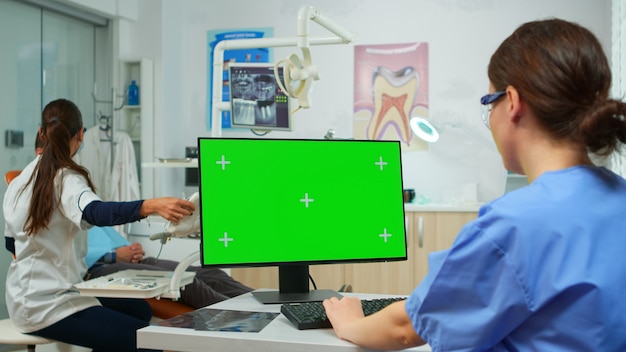 Enfermeira estomatologista olhando para o tablet de tela verde enquanto o dentista especialista examina o paciente com dor de dente sentado na cadeira de estomatologia. Mulher usando monitor com chroma key izolated pc key moc