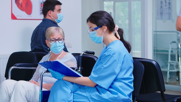 Enfermeira, explicando o diagnóstico a uma mulher idosa com deficiência em cadeira de rodas. Assistente usando máscara facial contra infecção por coronavírus na sala de espera do hospital.