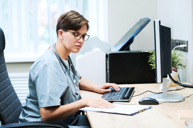 Enfermeira jovem concentrada em óculos sentado na mesa e usando computador e papéis ao preencher o relatório médico