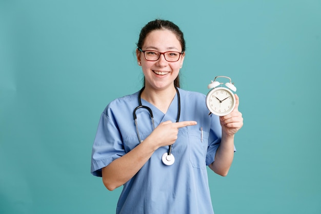 Enfermeira jovem de uniforme médico com estetoscópio no pescoço segurando o despertador apontando com o dedo indicador para ele feliz e positivo sorrindo alegremente sobre fundo azul