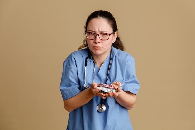 Enfermeira jovem de uniforme médico com estetoscópio no pescoço segurando pílulas olhando para a câmera confusa franzindo a testa em pé sobre fundo marrom